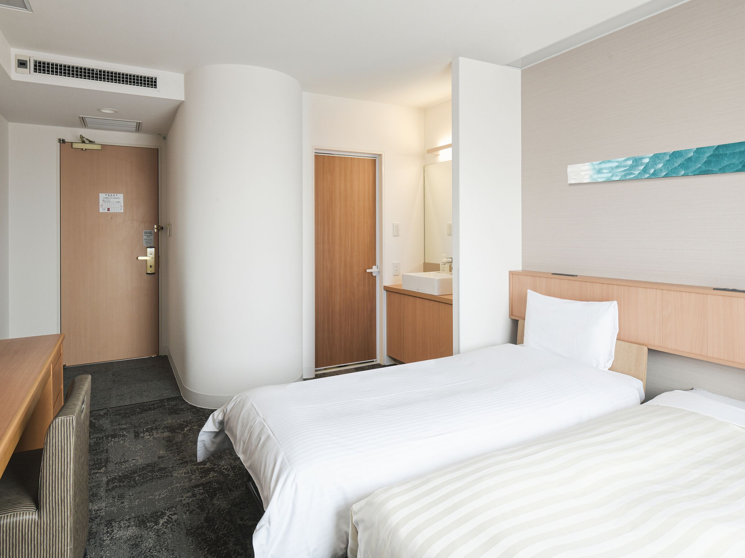 Habitación de hotel con dos camas, un escritorio de madera, un baño con lavamanos y espejo, y una obra de arte colgada en la pared.