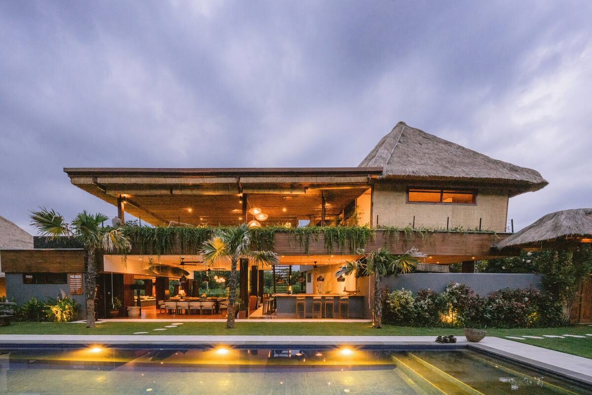 A modern two-story house with a pool, outdoor seating, and a thatched roof.