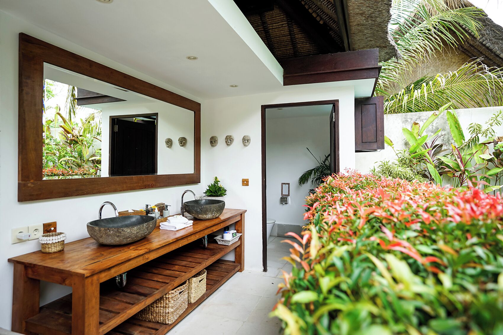 Un baño con tocador de madera, dos lavamanos de piedra, un espejo grande y vistas a un jardín tropical.