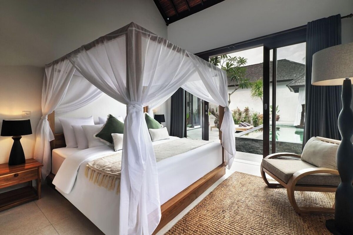 A bedroom with a four-poster bed, a nightstand with a lamp, a chair, and a view of a swimming pool and greenery outside.