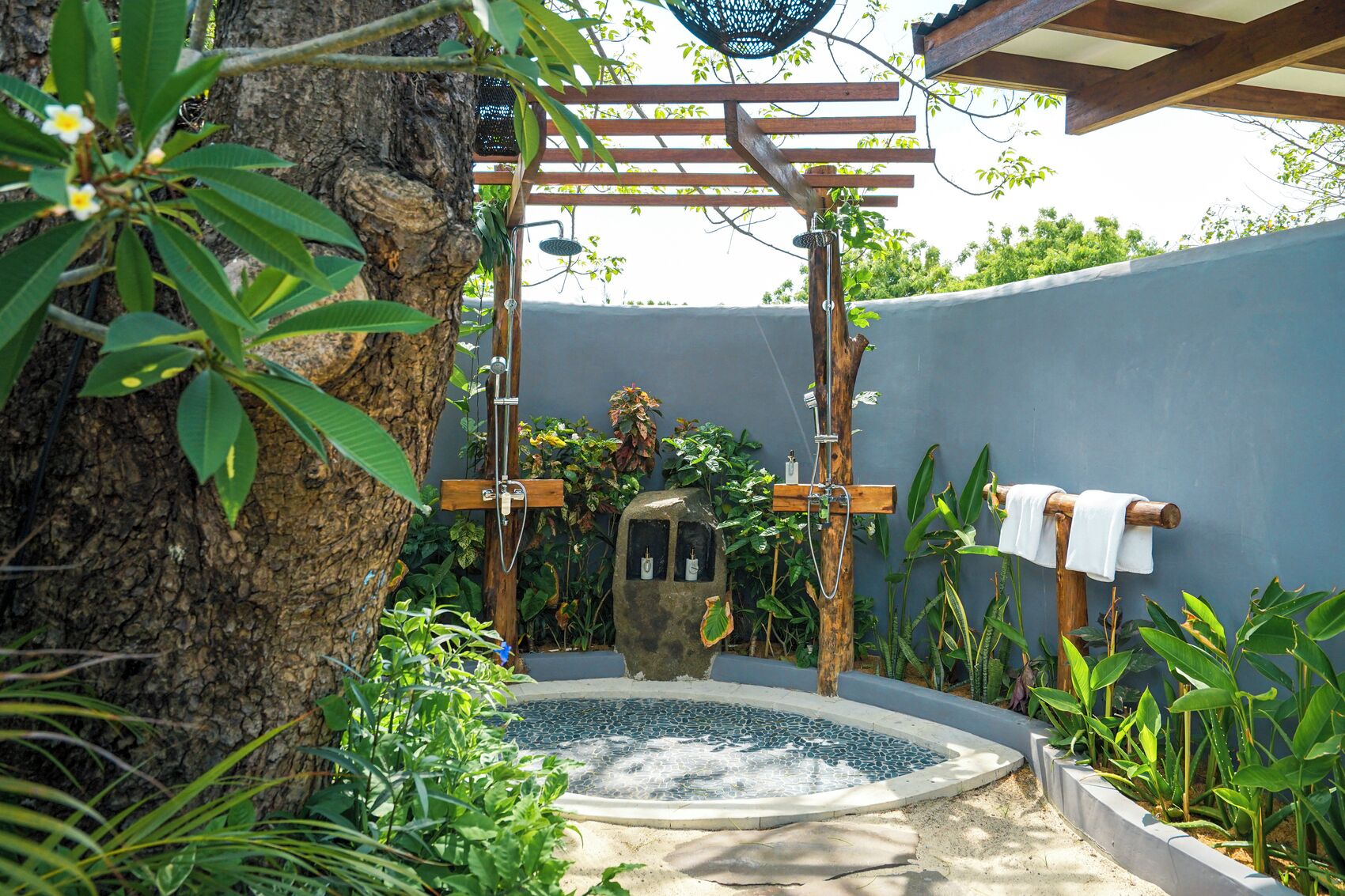 Un área de ducha privada al aire libre con un pérgola de madera, una pequeña estructura de piedra y un jacuzzi rodeado de exuberante vegetación.