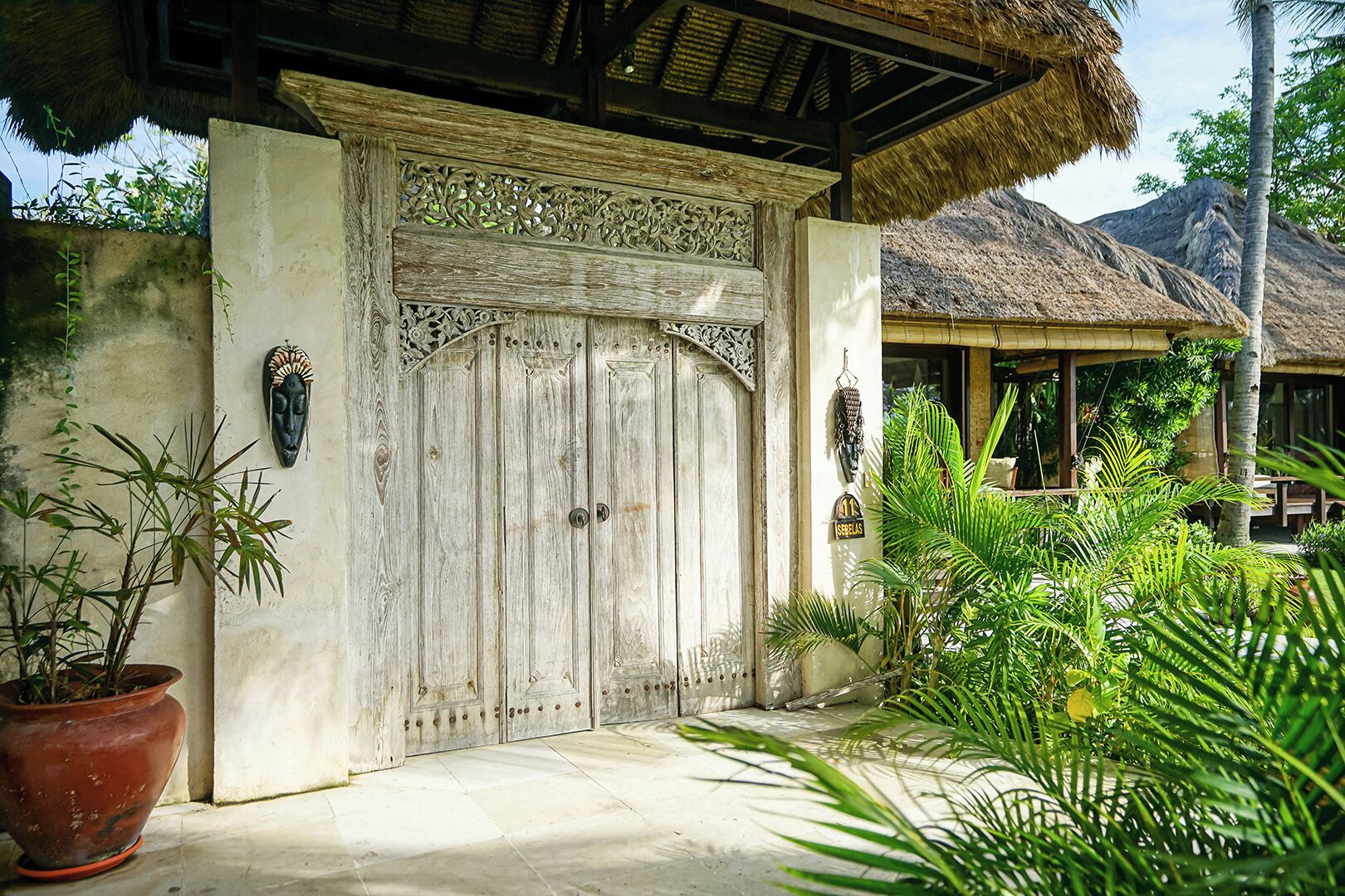A wooden gate with intricate carvings, a thatched roof structure, and tropical plants.