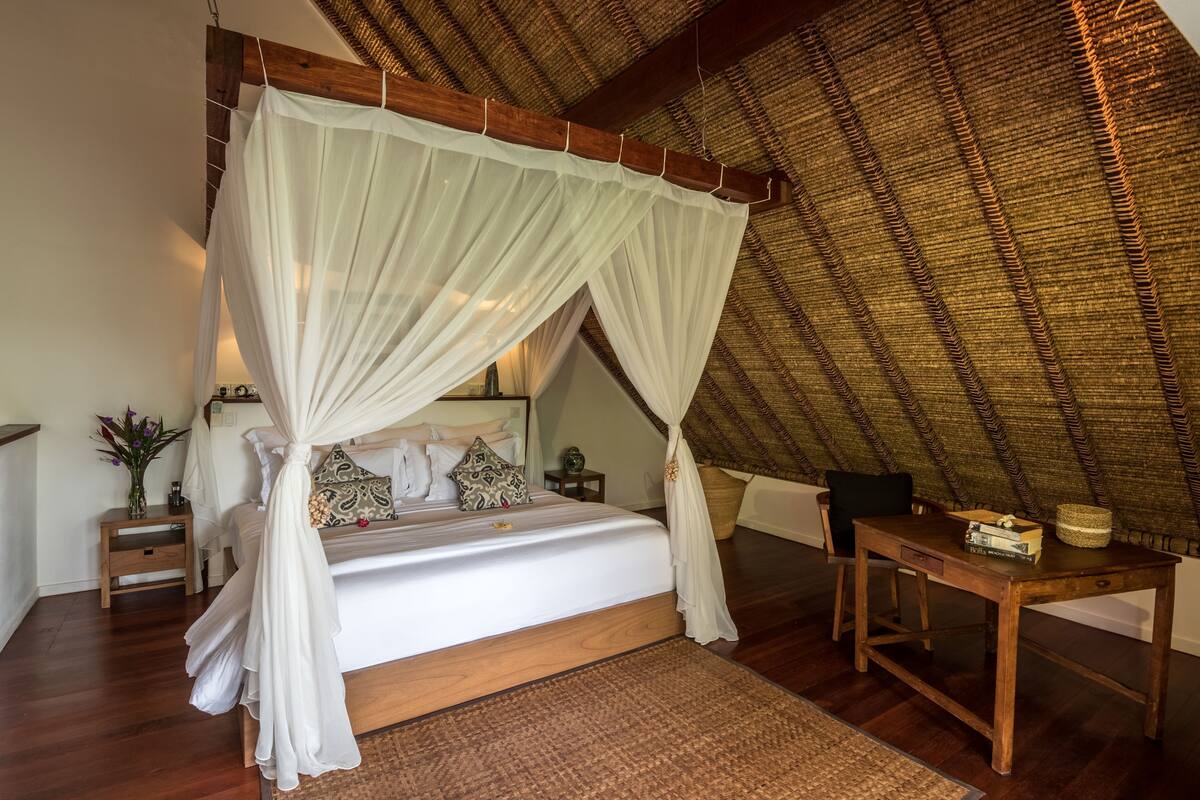 A bedroom with a canopy bed, wooden furniture, a woven ceiling, and a bedside table with a basket.