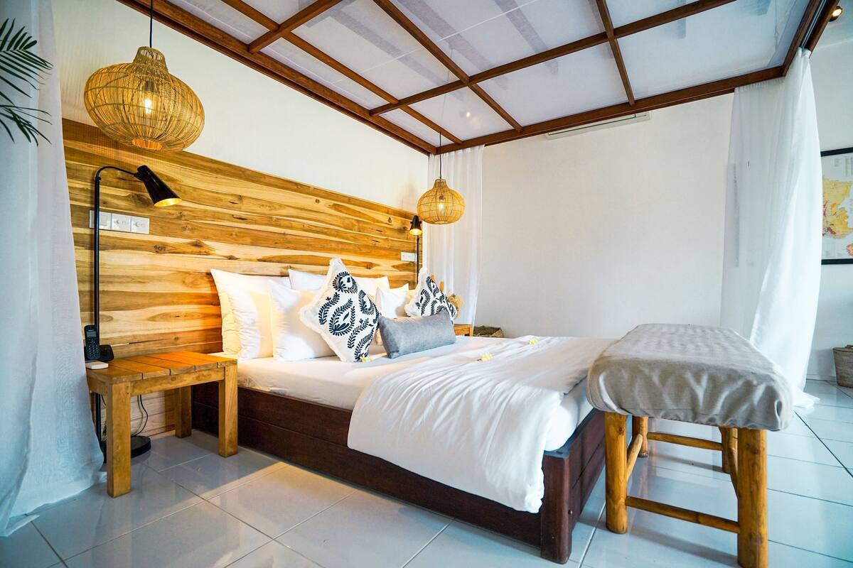 A bedroom with a wooden headboard, a bedside table, a lamp, a bed with white bedding, and a bench.