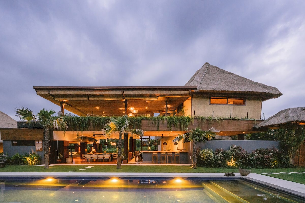 A modern two-story house with a pool, outdoor seating, and a thatched roof.