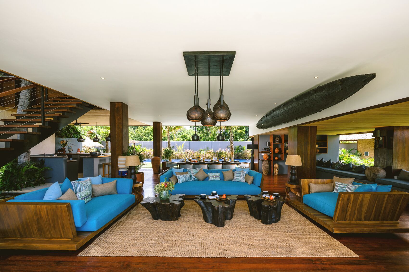 A spacious living area with a modern staircase, a large sofa set, and a dining area with a view of lush greenery.