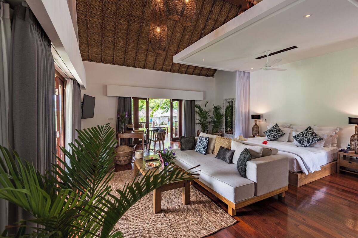 A modern hotel room with a large bed, a sofa, a dining area, and a view of the outdoors.