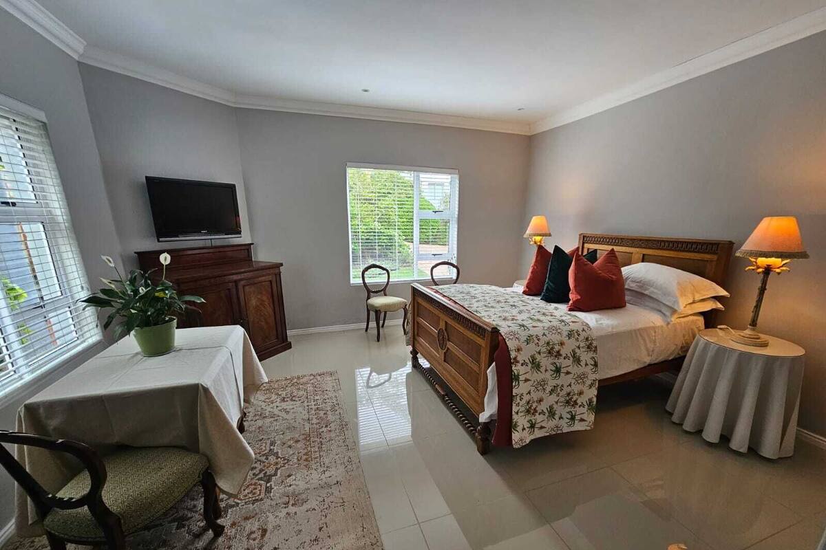 Un dormitorio con cama, lámparas de noche, televisor, silla, mesa y ventana.