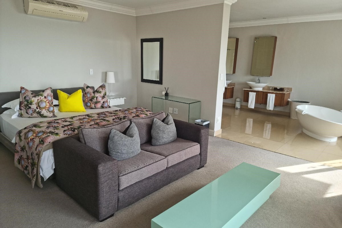 Una habitación de hotel moderna con una cama grande, un sofá gris, una mesa de centro de vidrio, un baño con bañera y un espejo.