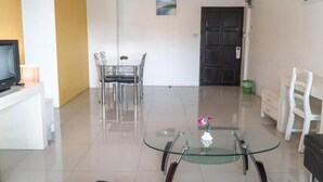 Una sala de estar moderna con una mesa de centro de vidrio, un televisor sobre un mueble blanco, una zona de comedor con una mesa y sillas, y una puerta negra.