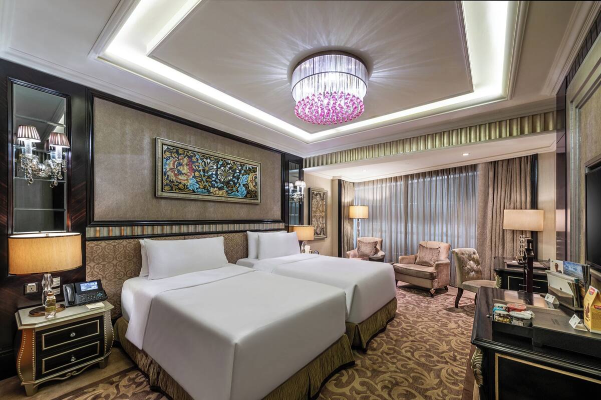 A hotel room with two beds, a chandelier, a painting, a desk with a lamp, and a chair.