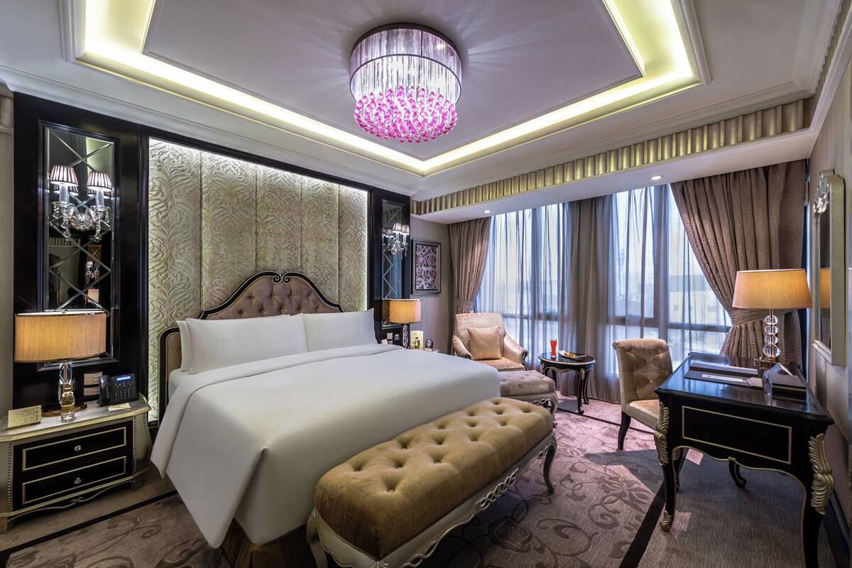 A spacious hotel room with a large bed, a tufted bench, a desk, and a chandelier.