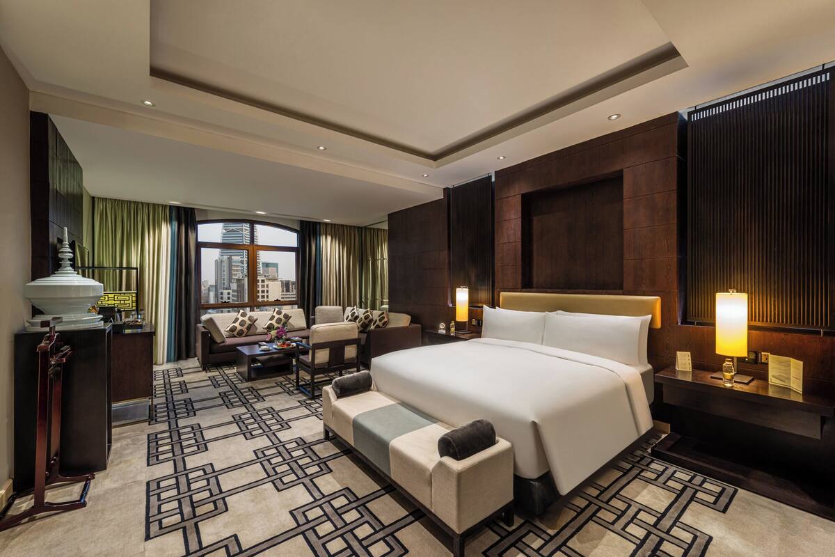 A modern hotel room with a large bed, a sofa, a desk, and a view of the cityscape.