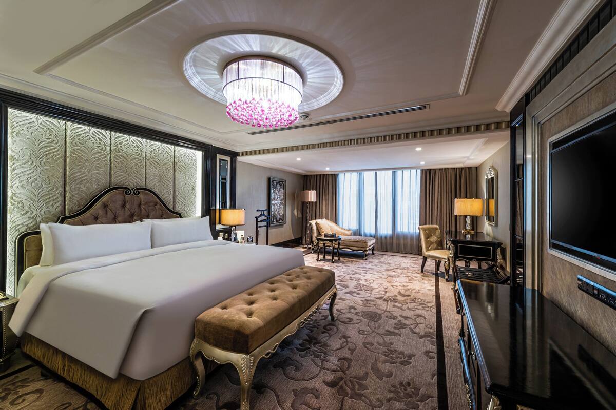 A spacious hotel room with a large bed, a sitting area with a chair and a table, a television, and a chandelier.