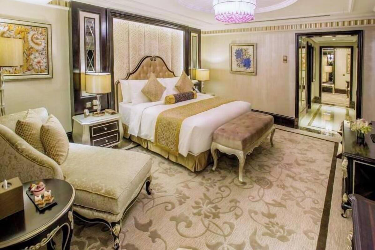 A hotel room with a large bed, a sitting area with a chair and ottoman, a nightstand, a mirror, and a chandelier.