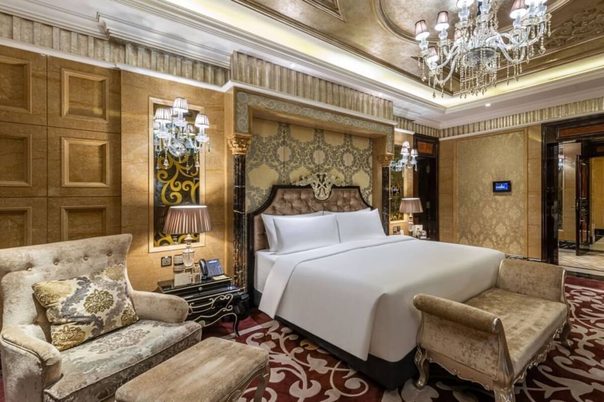 A luxurious hotel room with a large bed, two armchairs, a chandelier, and ornate wall decorations.