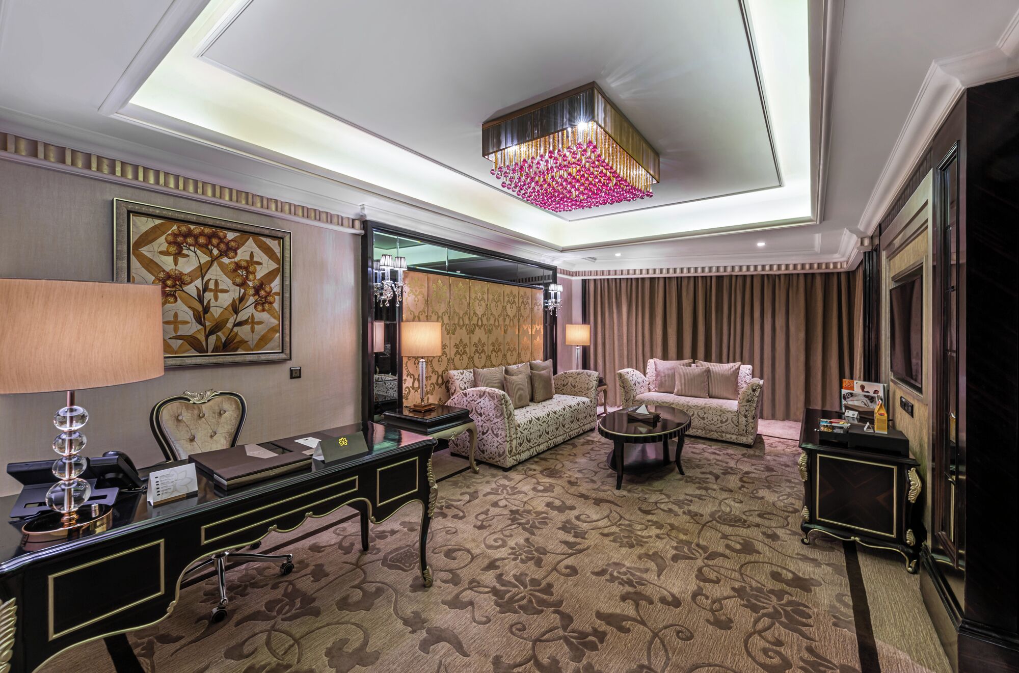 A luxurious hotel room with a large desk, two sofas, a chandelier, and a patterned carpet.