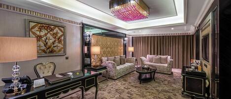 A luxurious hotel room with a large desk, two sofas, a chandelier, and a patterned carpet.