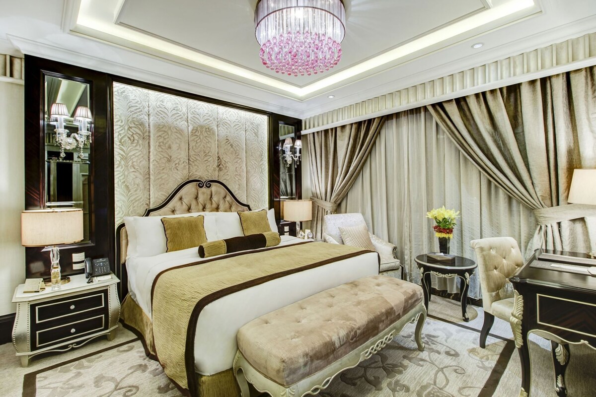 A luxurious hotel room with a large bed, a sitting area with a piano, a desk, and a chandelier.