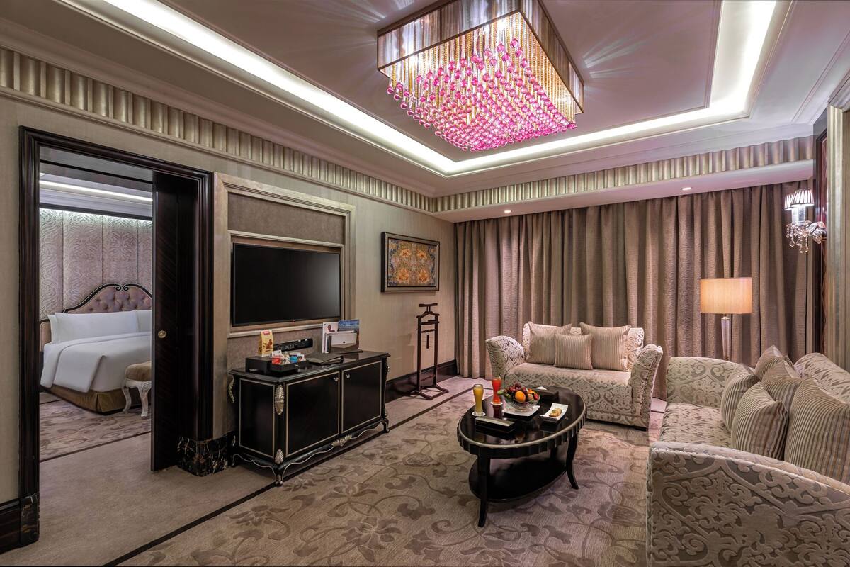 A luxurious hotel room with a large bed, a flat-screen TV, a dark wooden console table, a patterned sofa set, and a decorative chandelier.