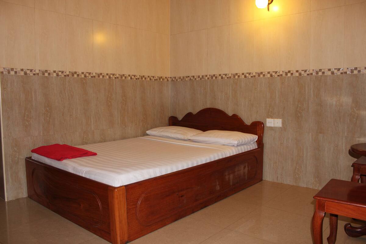 A wooden bed with white linens and a red blanket, a small wooden table, and a wall with a patterned border.