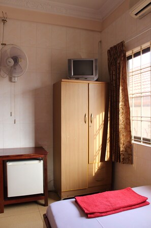 Een kamer met een bed, een houten garderobe, een kleine witte koelkast, een ventilator aan de muur en een raam met gordijnen.