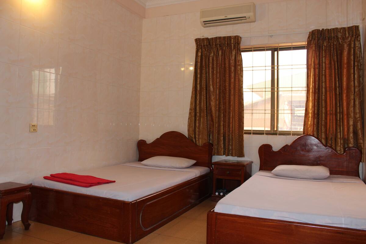 Habitación de hotel con dos camas de madera, una mesita de noche, una ventana con cortinas y un equipo de aire acondicionado.