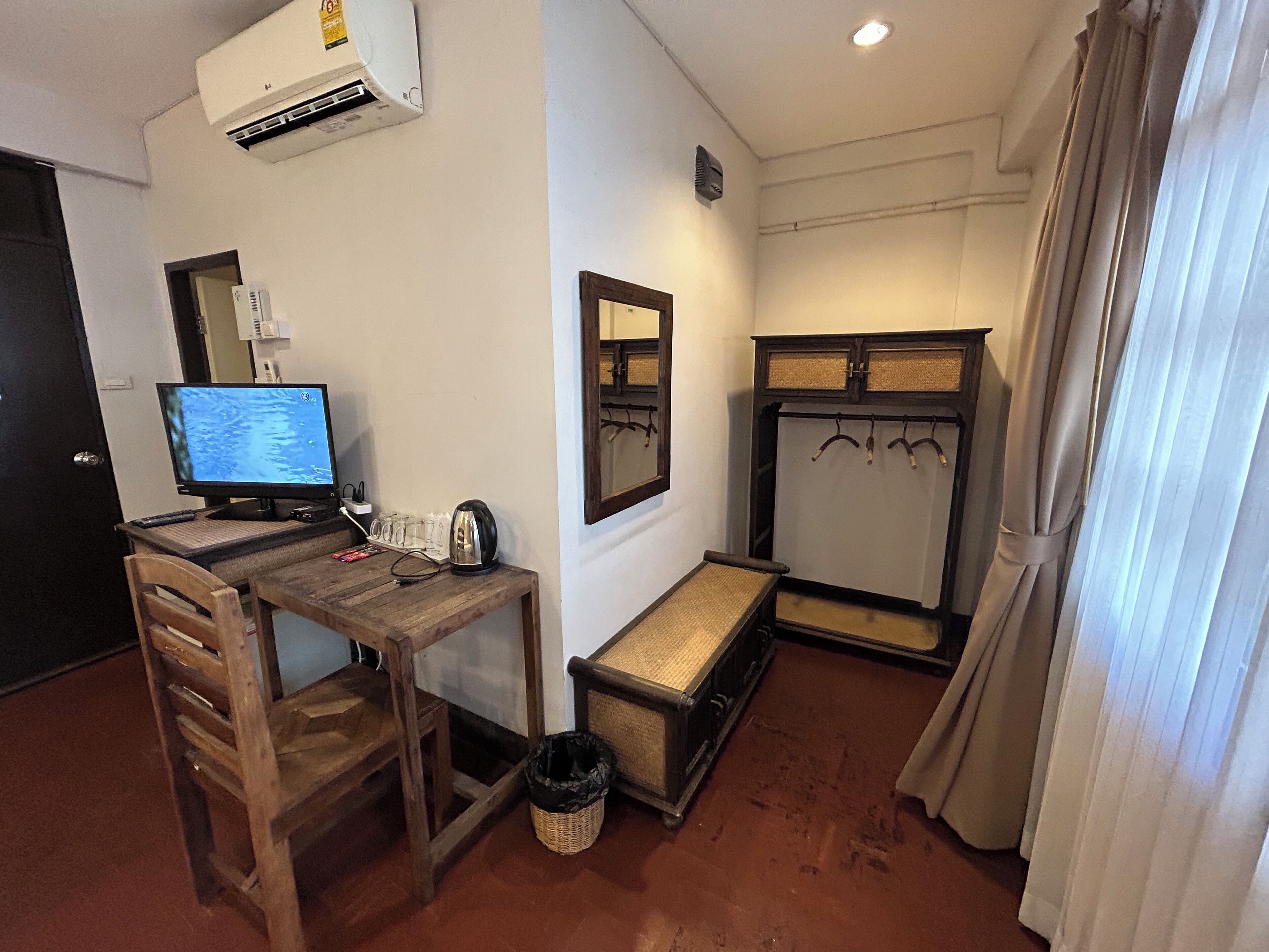 Een compacte kamer met een houten bureau, een computer, een waterkoker, een klein bankje, een spiegel en een garderobe.