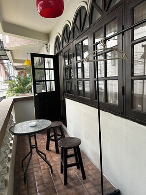 Un balcon avec une table ronde et deux tabourets, une lampe suspendue rouge et une fenêtre avec un cadre en métal.