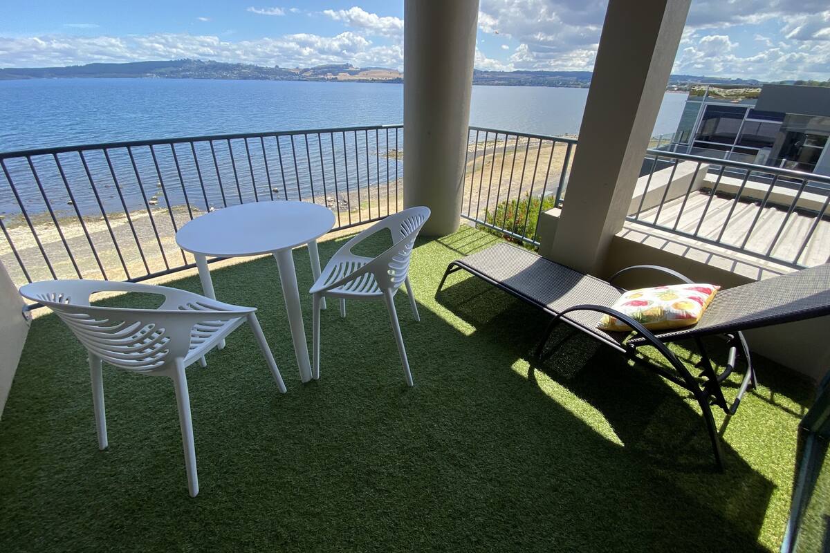 A balcony with a view of the sea, a round table, two white chairs, and a lounge chair with a cushion.