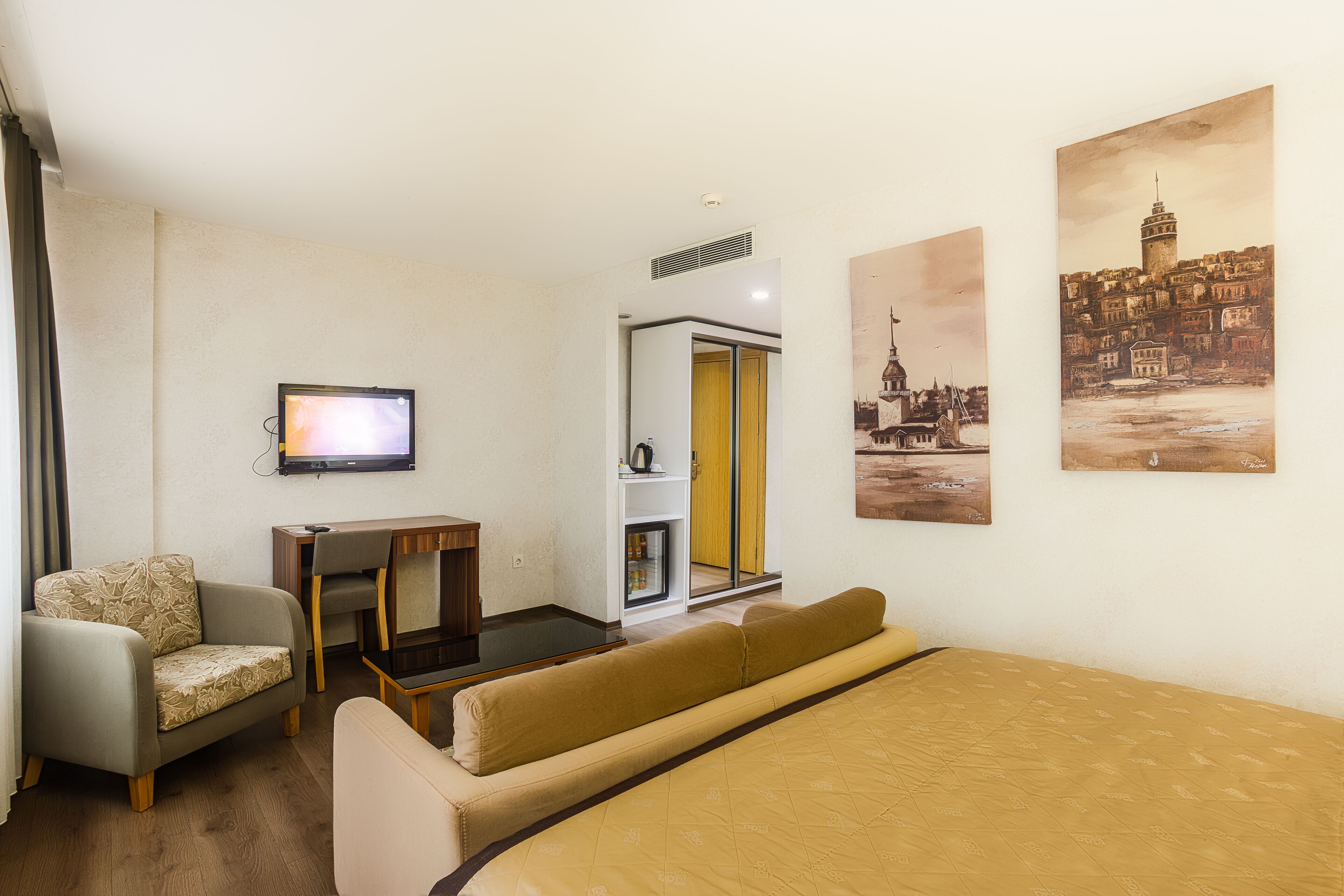 A hotel room with a bed, a sofa, a desk, a television, and artwork on the walls.