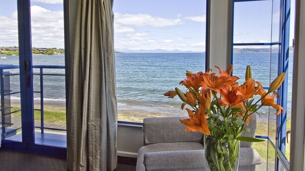 Una habitación con un amplio ventanal que ofrece vistas al mar y a las montañas, un sofá gris y un jarrón con flores naranjas.