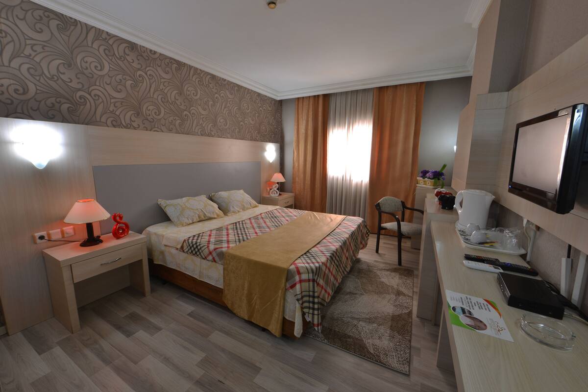 A hotel room with a bed, bedside tables, a desk, a chair, a TV, and a window with curtains.