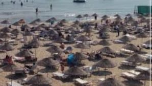 Una playa con numerosos paraguas y tumbonas, barcos en el agua y un cielo despejado.