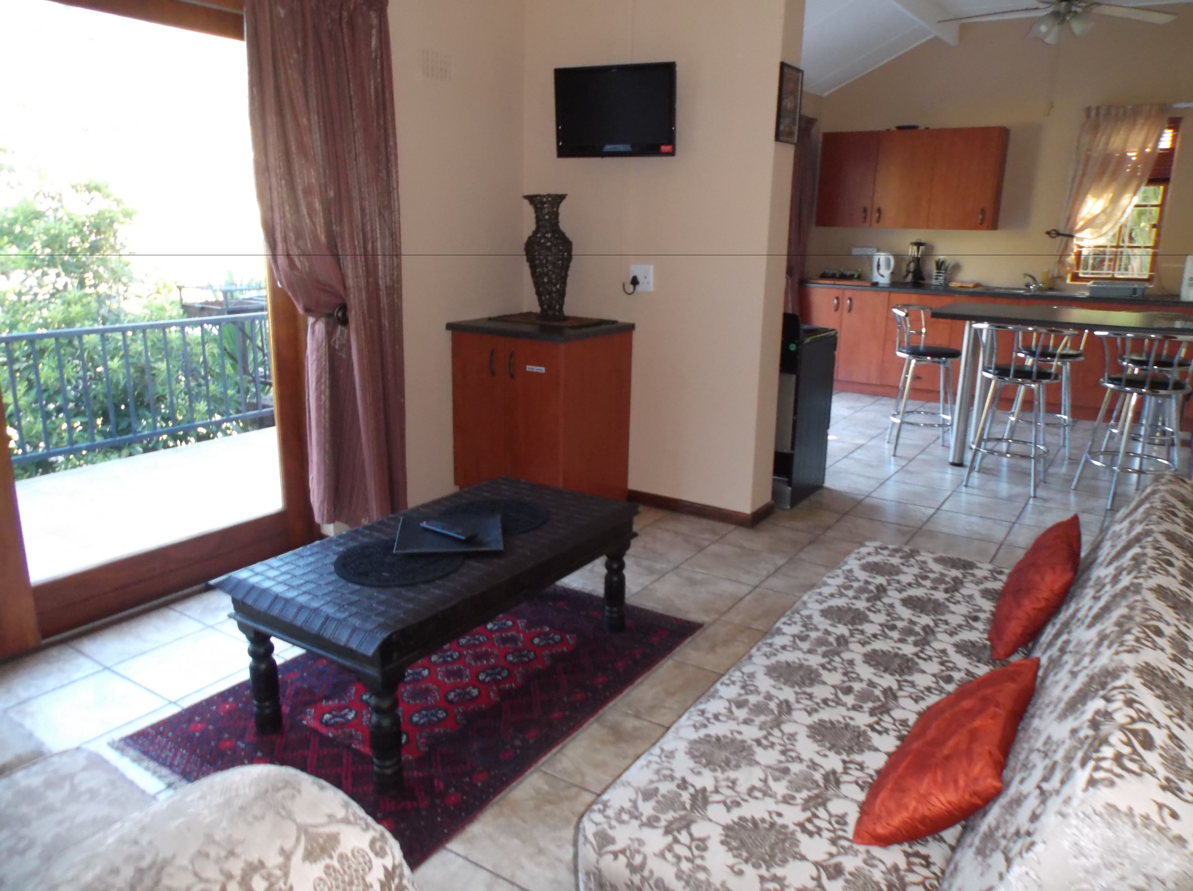Un salon comprenant un canapé, une table basse et un téléviseur fixé au mur. On aperçoit à l’arrière-plan un coin cuisine avec un bar et des chaises.