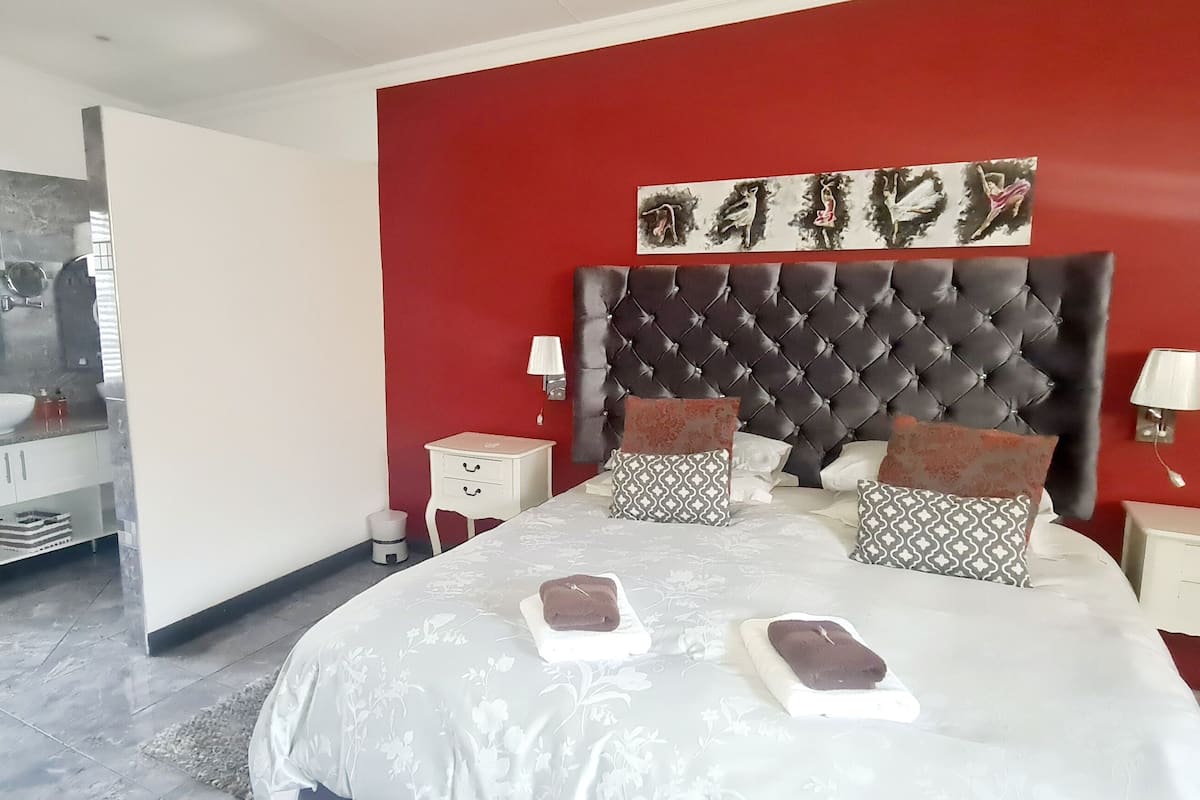 Een moderne slaapkamer met een rode accentmuur, een gestoffeerd hoofdeinde, een wit bed met bloemenbeddengoed en een nachtkastje met een lamp.