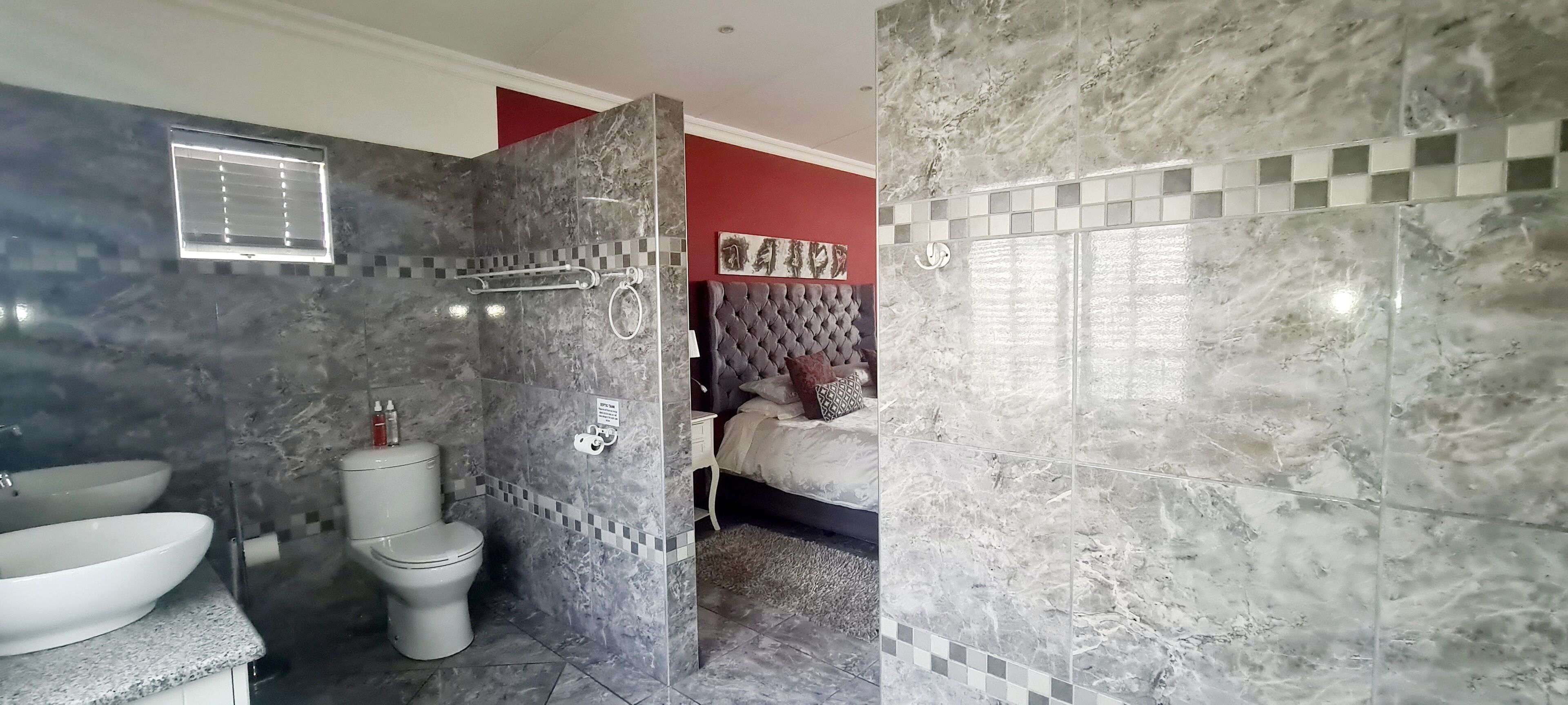 Une salle de bain avec douche, toilettes et lavabo, des murs en marbre et un lit dans la pièce adjacente.