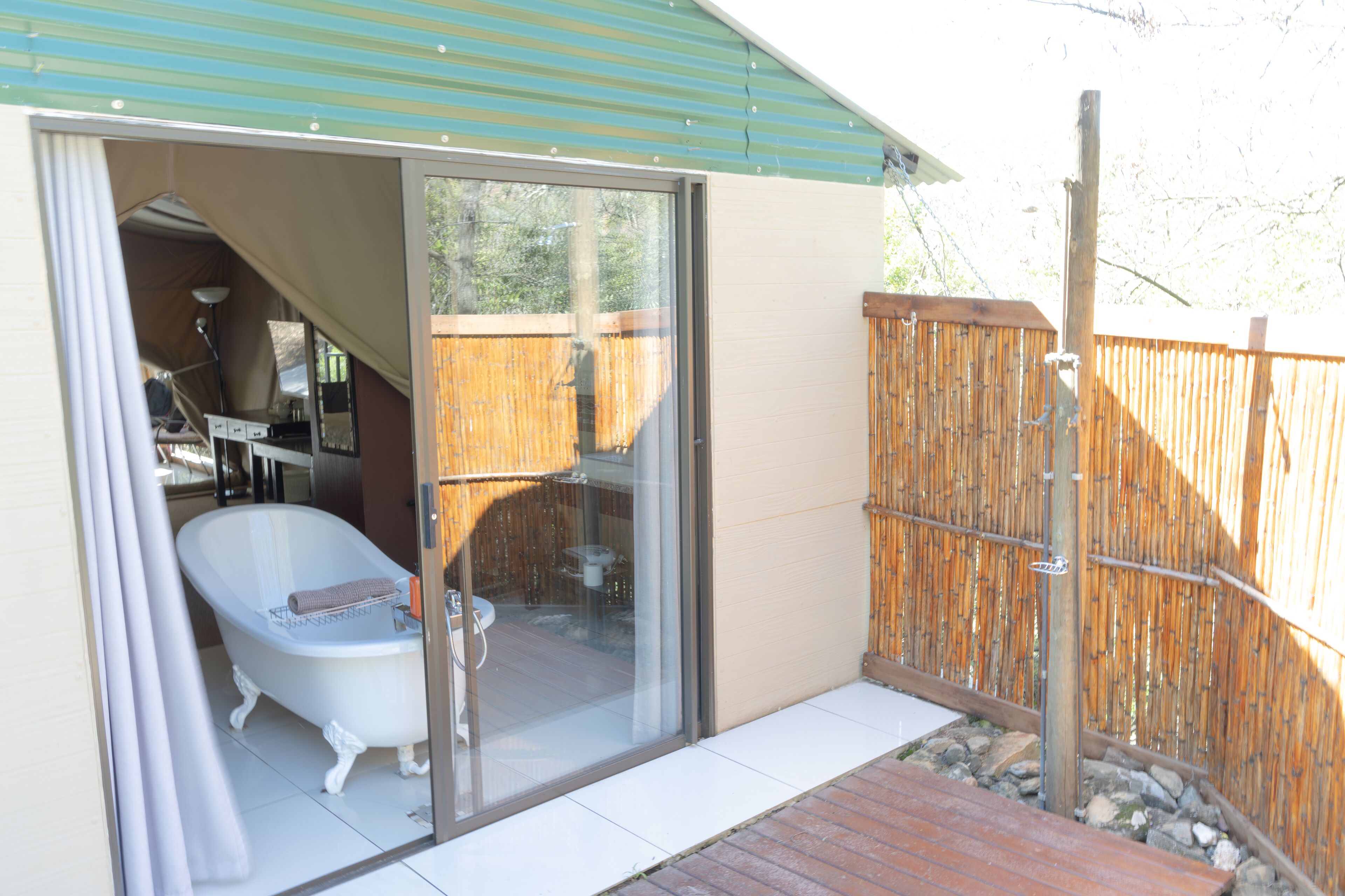 Un baño con una bañera de patas de león blanca, una puerta de cristal que da a un patio, y una valla de bambú al fondo.