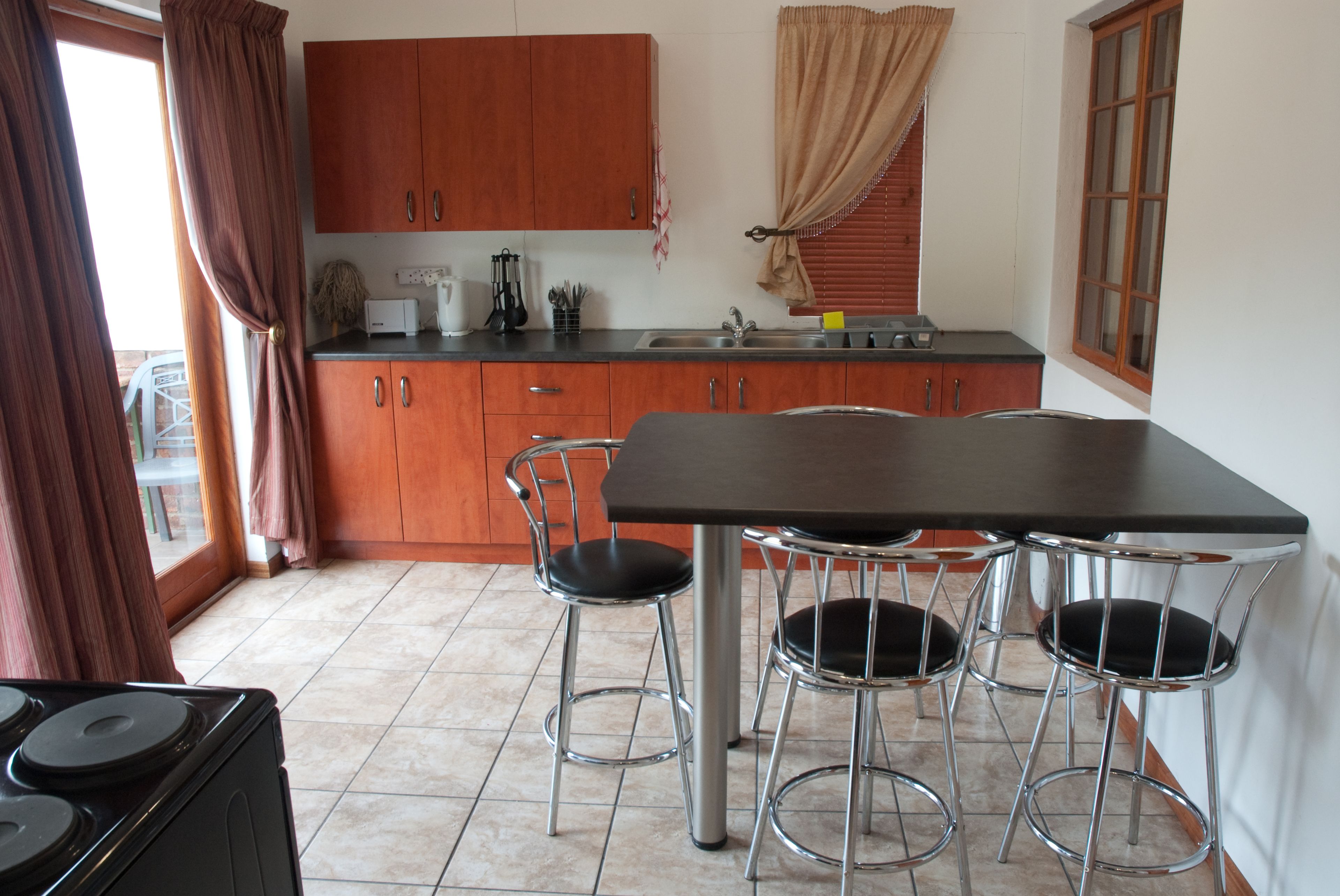 Uma cozinha com mesa de jantar e quatro banquetas altas, armários de madeira, uma janela com cortinas e um fogão.
