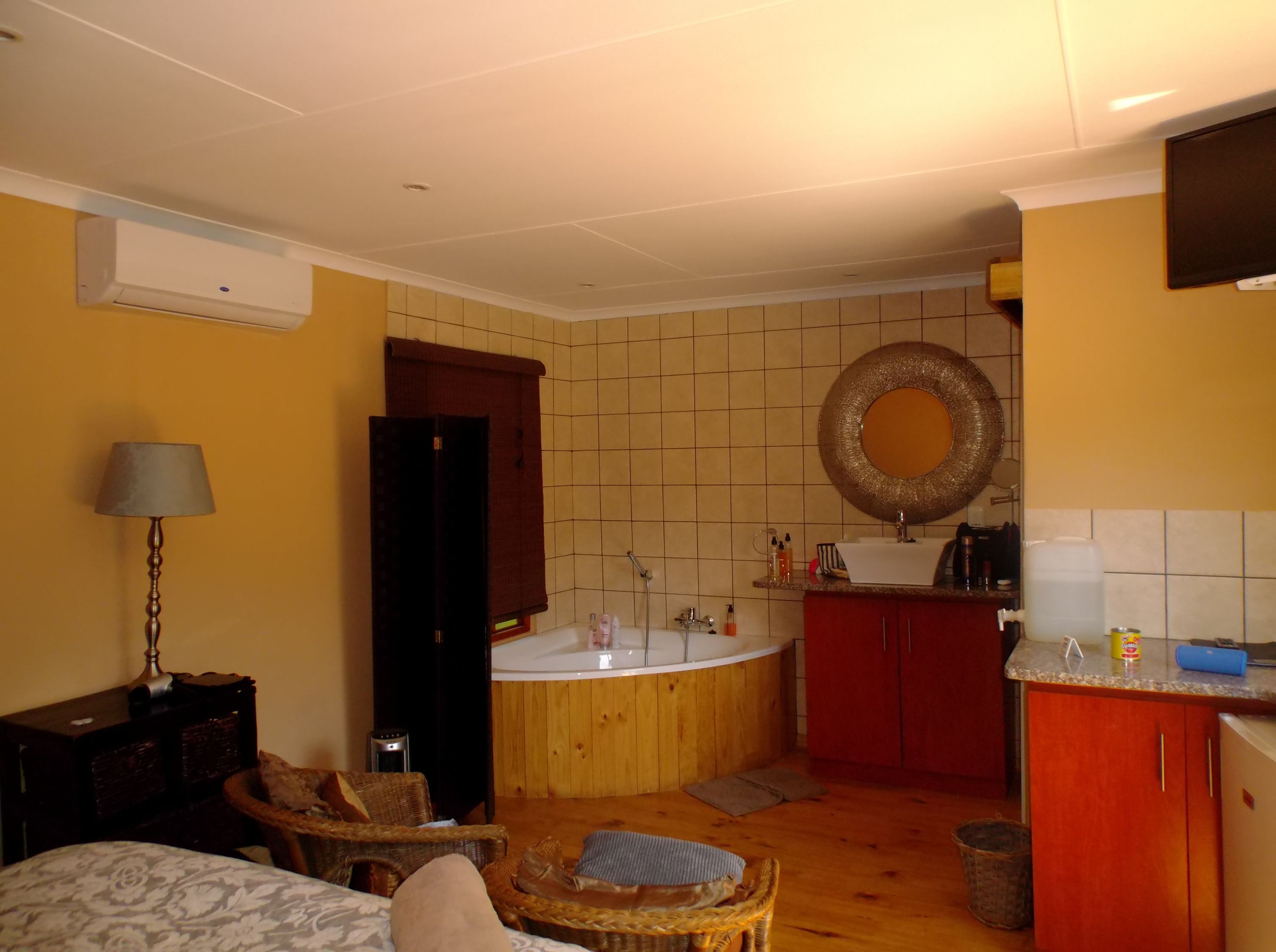 A compact room with a bed, a small table with a lamp, a wicker chair, a wooden cabinet, a round mirror, a sink, and a bathtub.