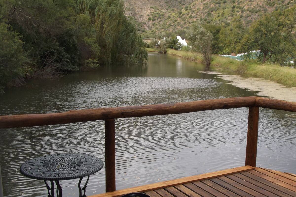 Een houten terras met een metalen tafel en bank, uitkijkend over een rustige rivier, omringd door groen en heuvels.