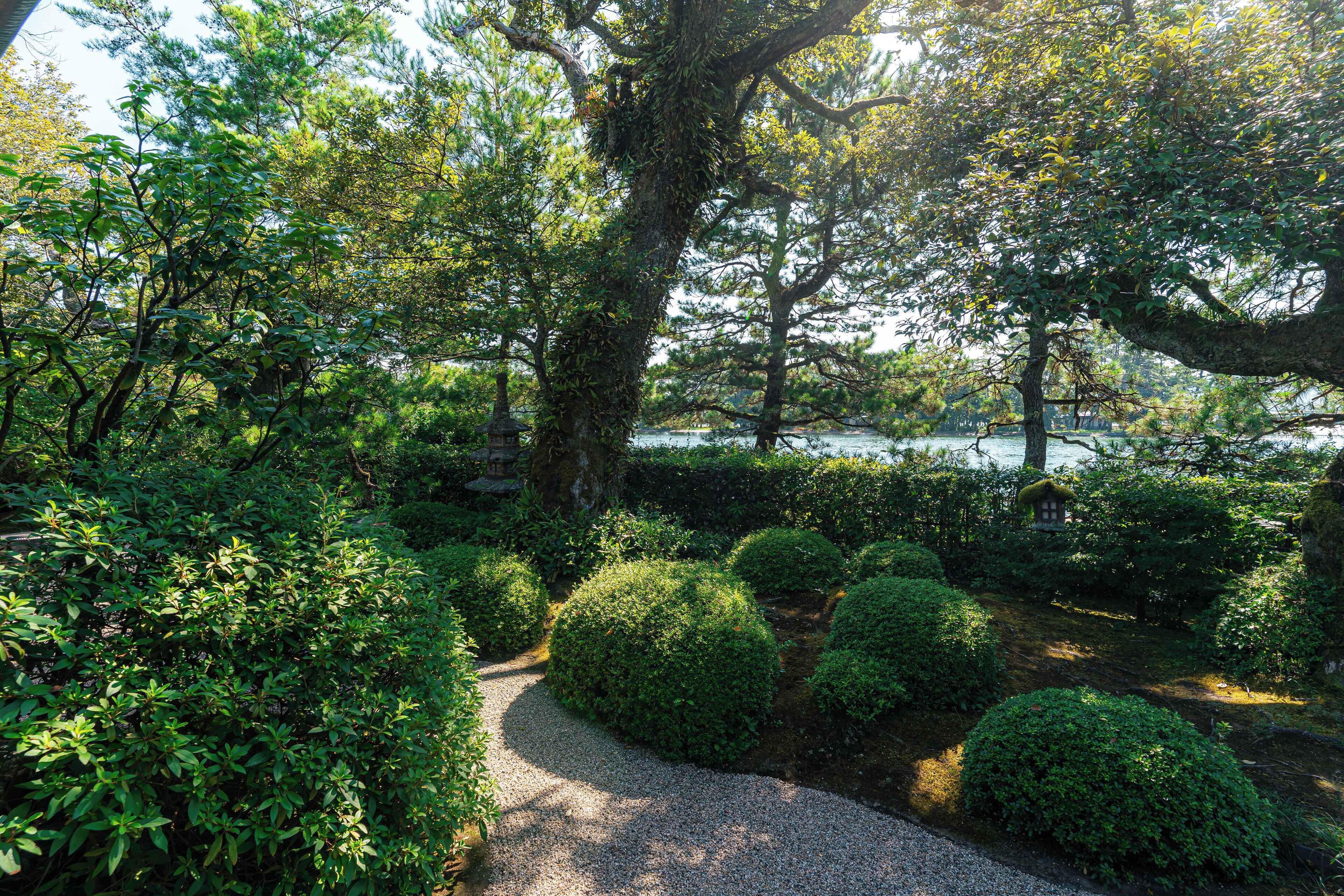 A serene garden with a gravel path, neatly trimmed bushes, and a small traditional building.