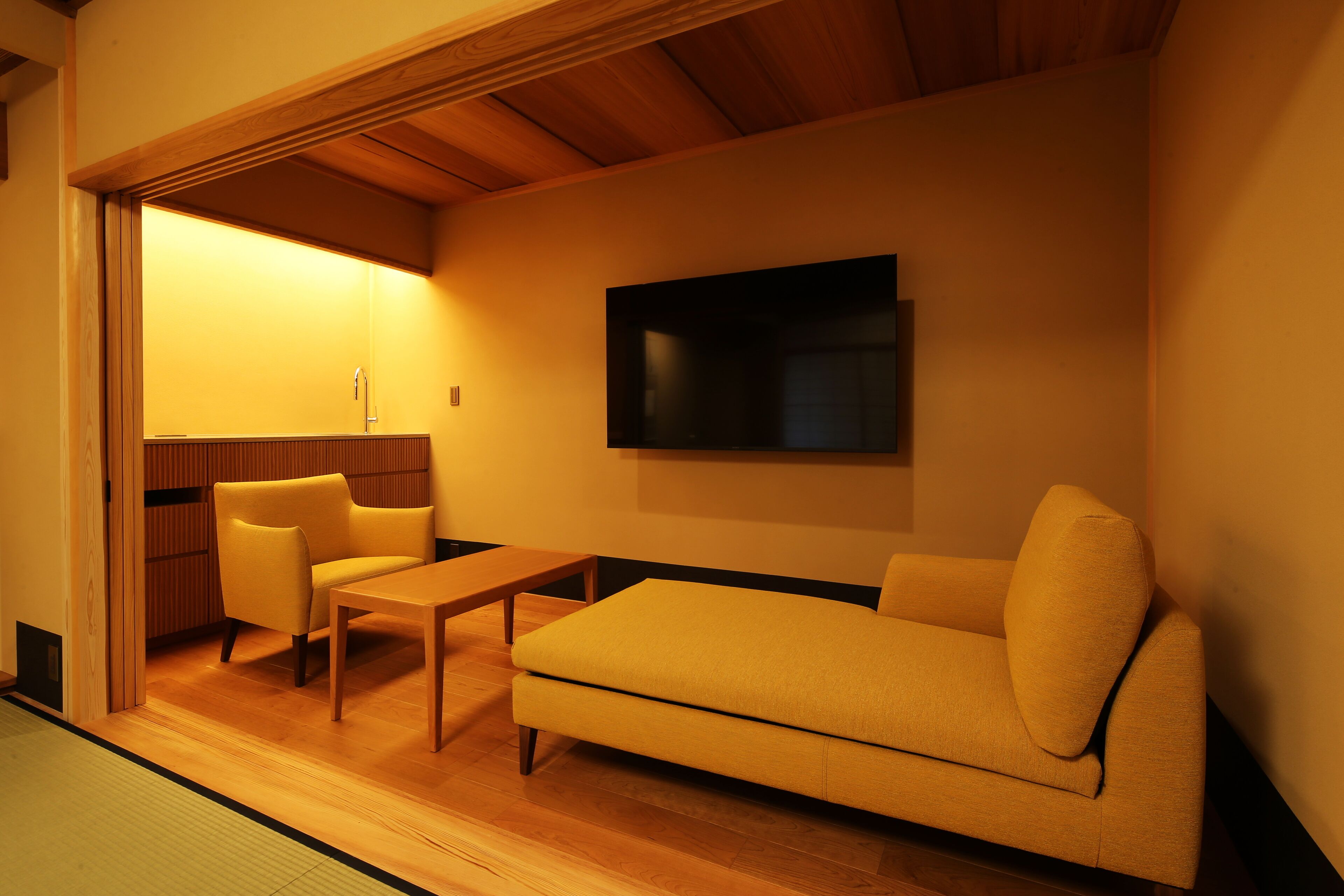 A modern living room with a sofa, a small table, and a chair. There is a wooden floor and a wall-mounted TV.