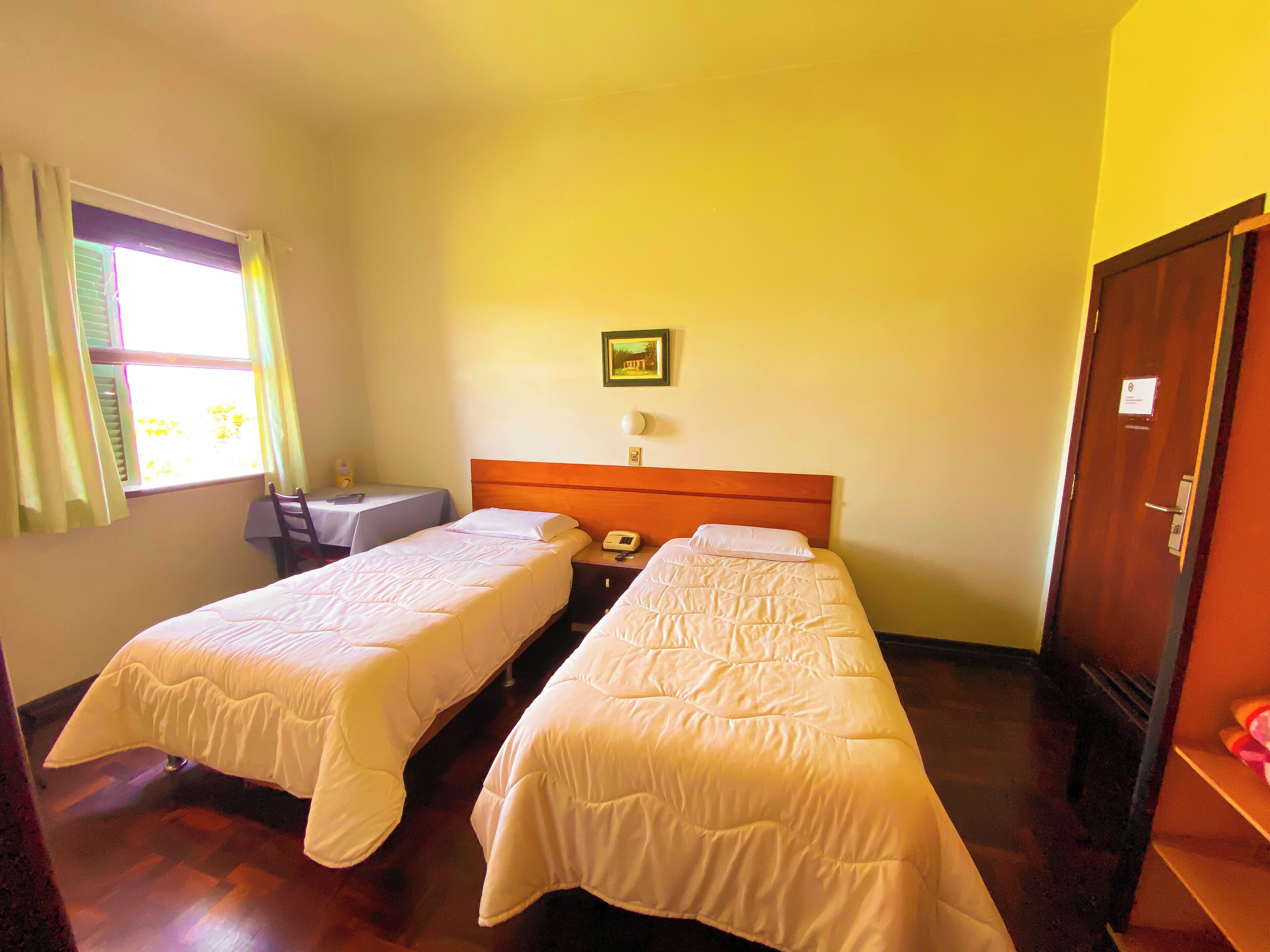 Habitación de hotel con dos camas individuales, una mesita pequeña, una silla, teléfono en la mesita de noche y una ventana con cortinas.