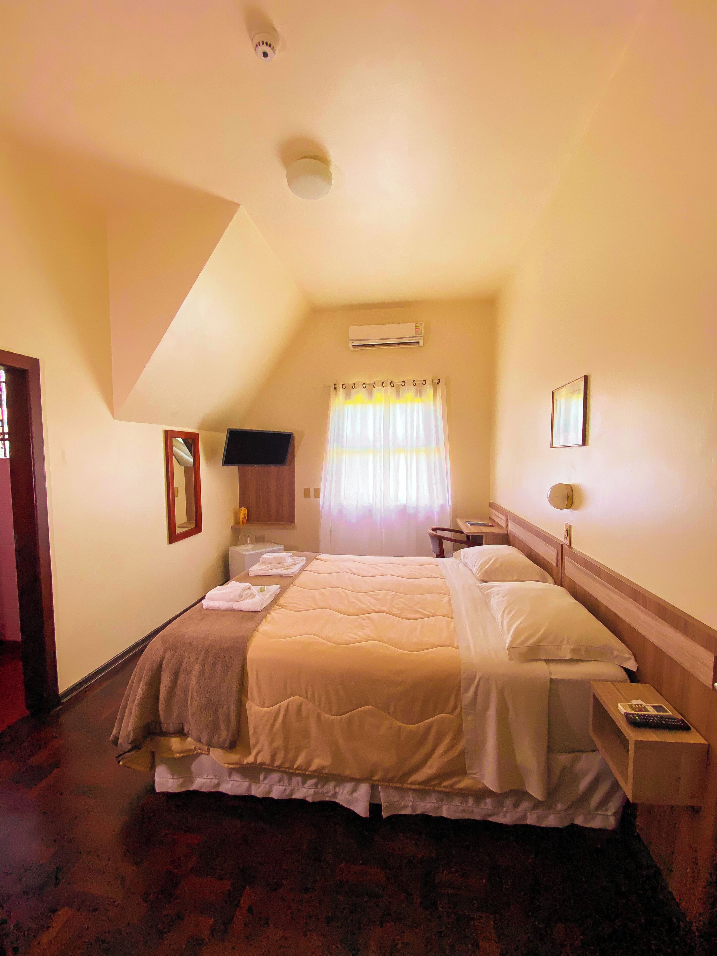 Habitación de hotel con cama, mesita de noche con control remoto, espejo, equipo de aire acondicionado y ventana con cortinas.