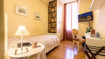 Un dormitorio con cama, mesita de noche, lámpara, estantería, silla, escritorio y televisor montado en la pared.