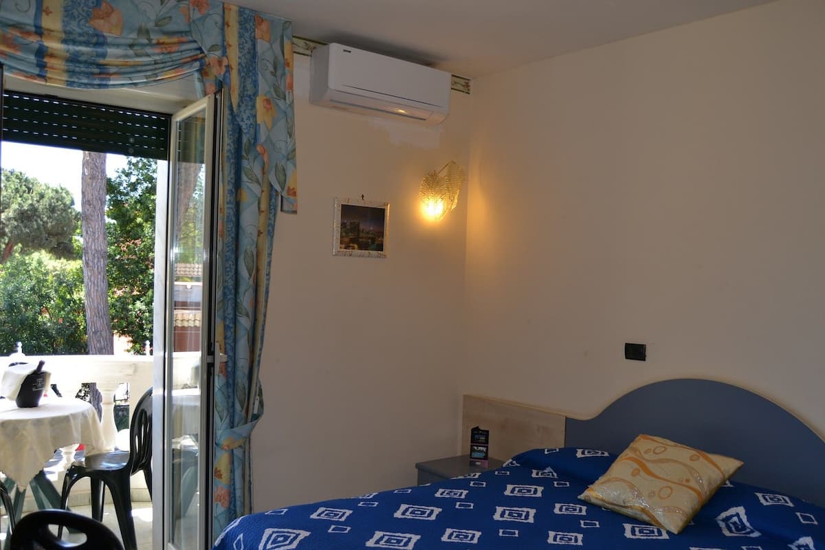 Un dormitorio con cama, mesita de noche, aire acondicionado instalado en la pared, ventana con cortinas y vistas al exterior.
