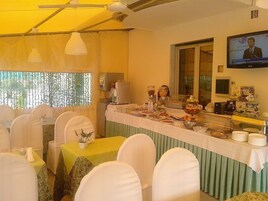Un comedor con mesas y sillas, una barra de buffet con alimentos, una máquina de café y una pantalla de televisión que muestra un noticiero.