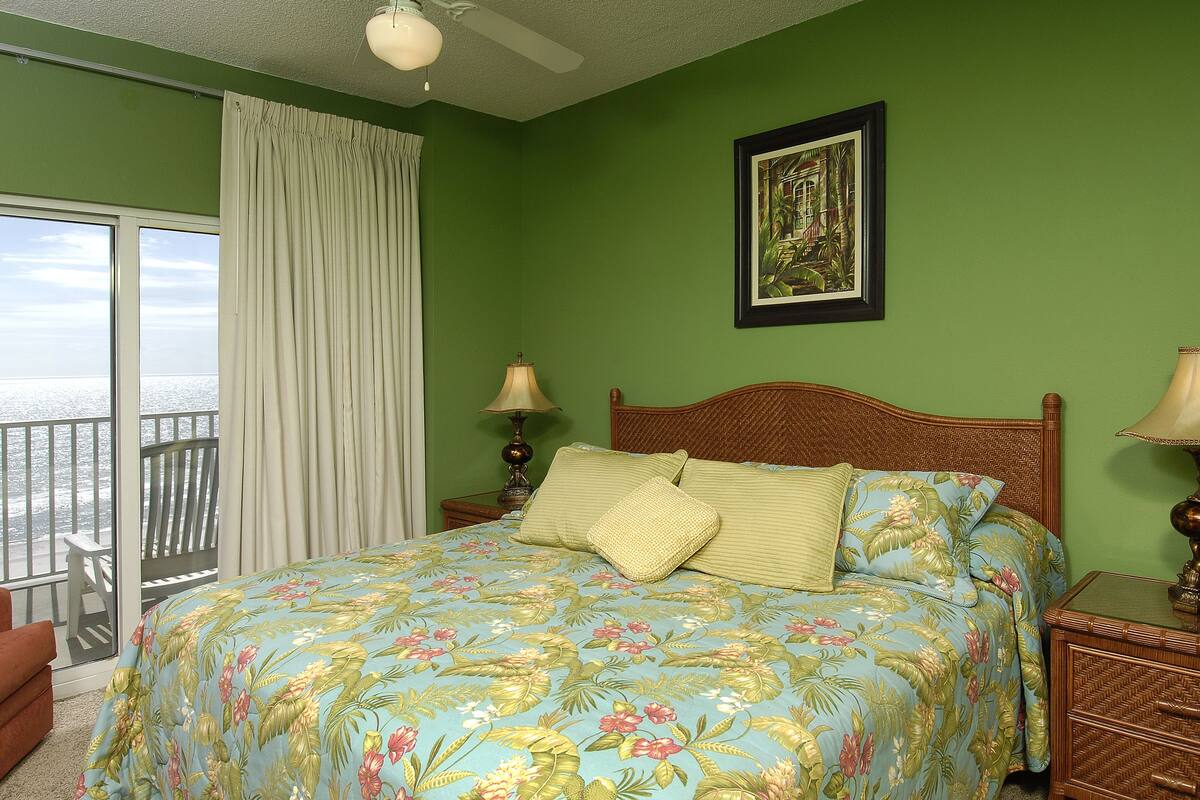 A bedroom with a large bed, a nightstand, a chair, a window with curtains, and a ceiling fan.