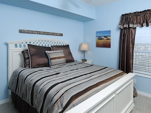 A bedroom with a bed, a nightstand, a lamp, a painting, and a window with curtains. - Tidewater By Wyndham Vacation Rentals (Orange Beach)